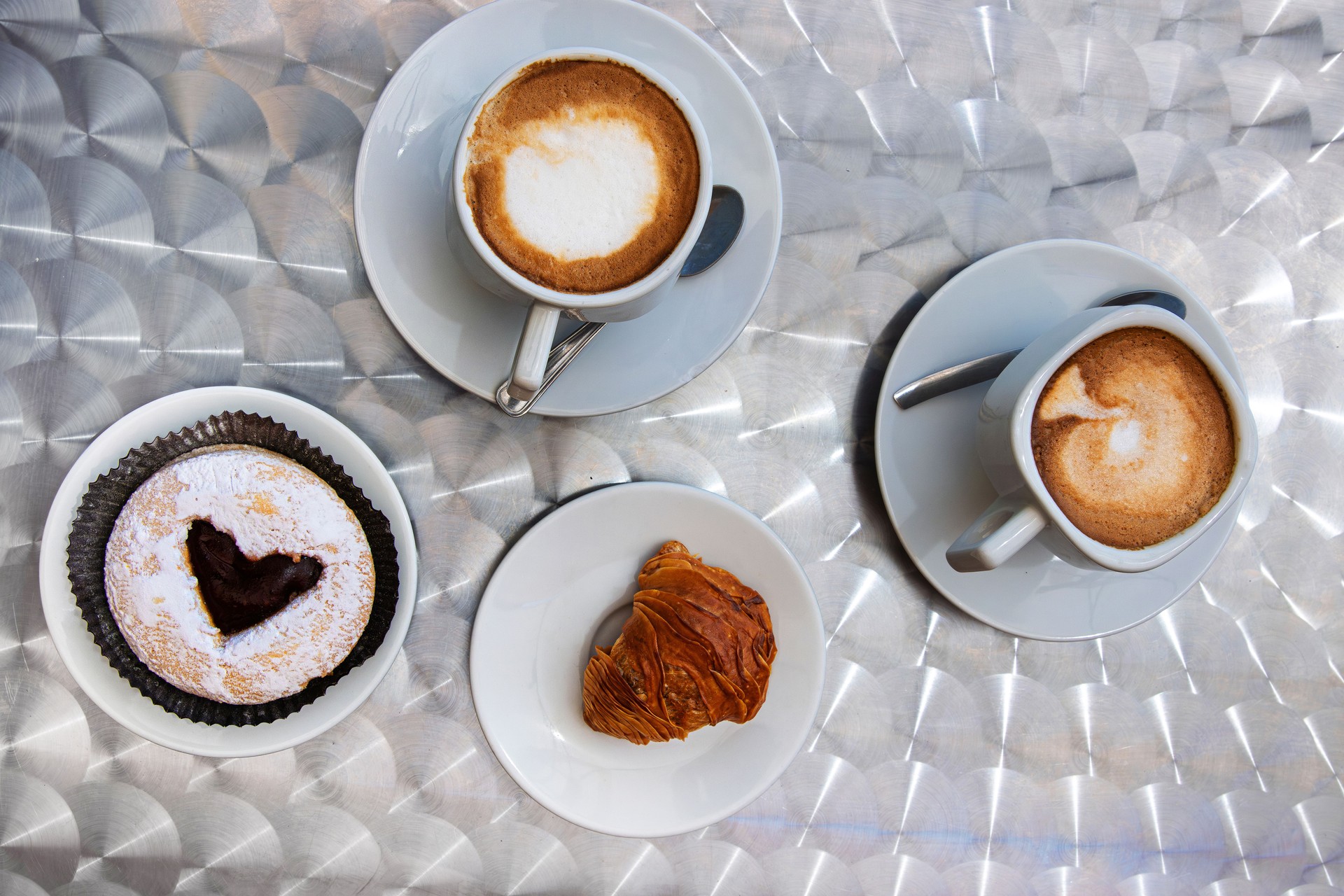 Overhead view of Italian breakfast with two cappuccinos, a chocolate filled pastry, and a  sfogliatelle on white dishes atop a textured metallic table.